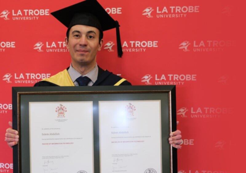 Salem is wearing his graduation clothes and cap while holding his degree in front of a red background