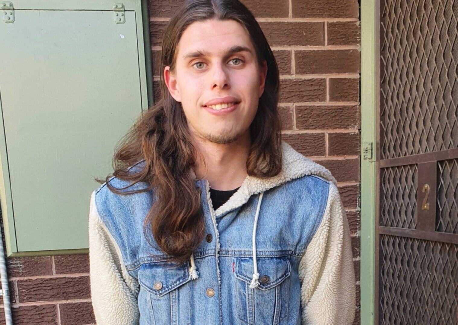 Benjamin is smiling looking at the camera. He is wearing his long hair out, and a blue denim hoodie.