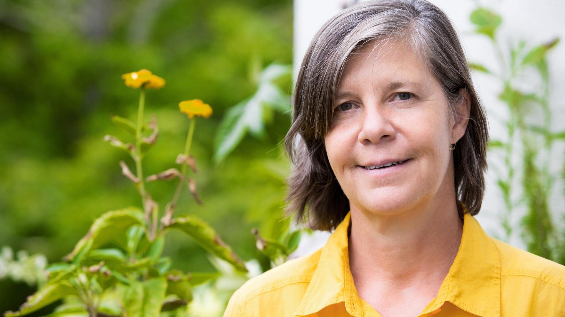 Woman wearing a yellow polo shirt is smiling at the camera. She is standing in a garden.