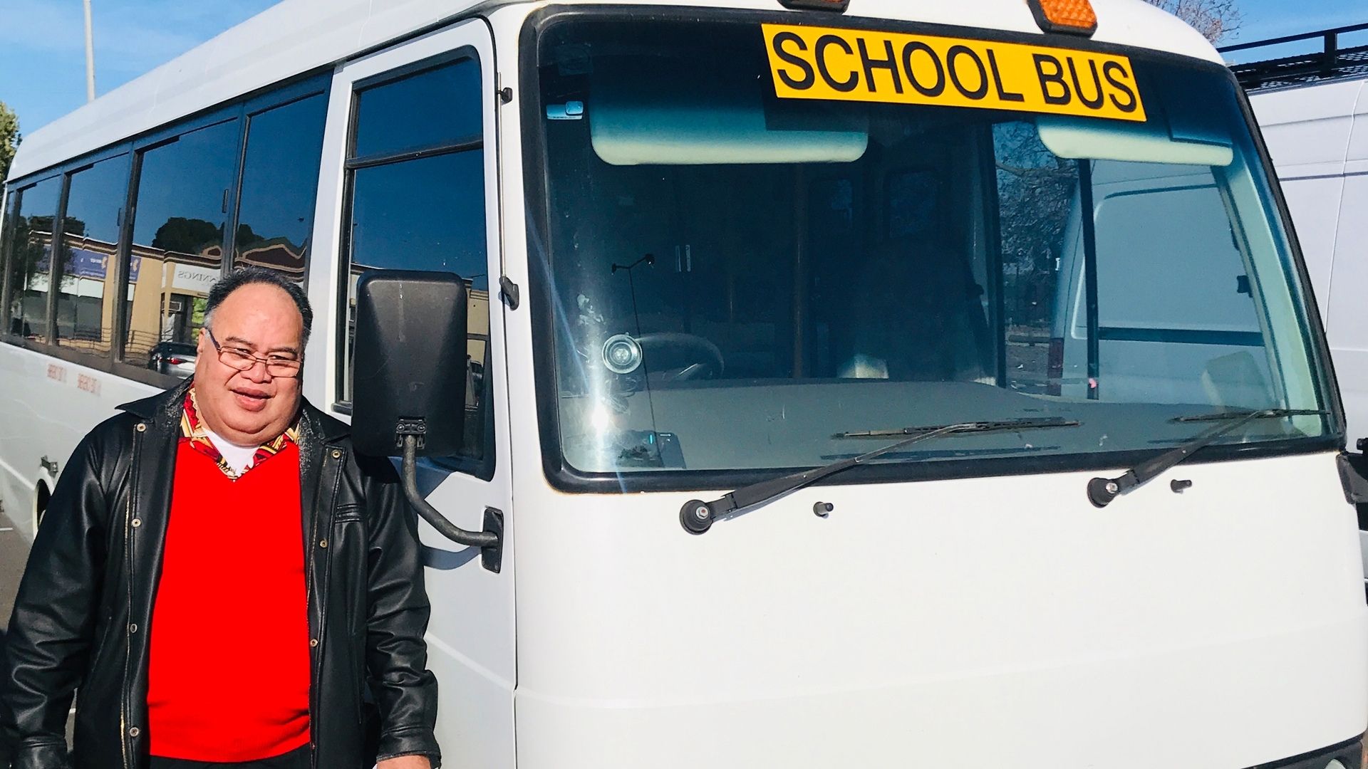 Fea stands beside the bus he drives. He is wearing a bright red jumper and smiling at the camera