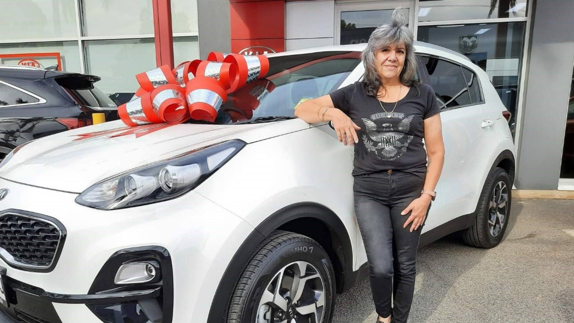 Christine is leaning on a white four wheel drive that has a big red bow on it. She is wearing a black t-shirt with jeans.