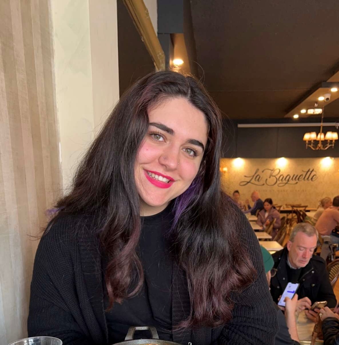 Lilith is sitting in a cafe leaning against a wall and smiling at the camera.