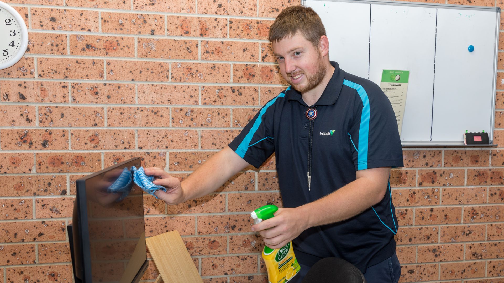 Daniel is cleaning a computer screen with spray and a wipe cloth. He is wearing his work uniform.