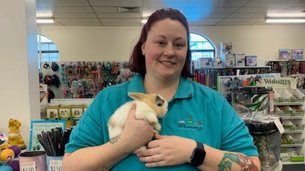 Rebecca, wearing her uniform, stands in a pet shop cuddling a white rabbit and smiling