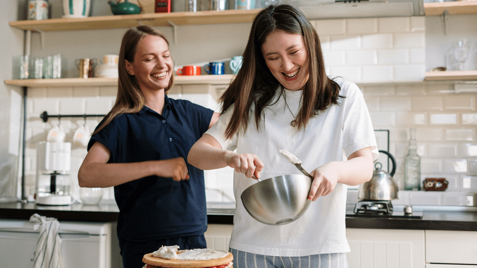 Young woman teaching another young woman how to frost a cake.