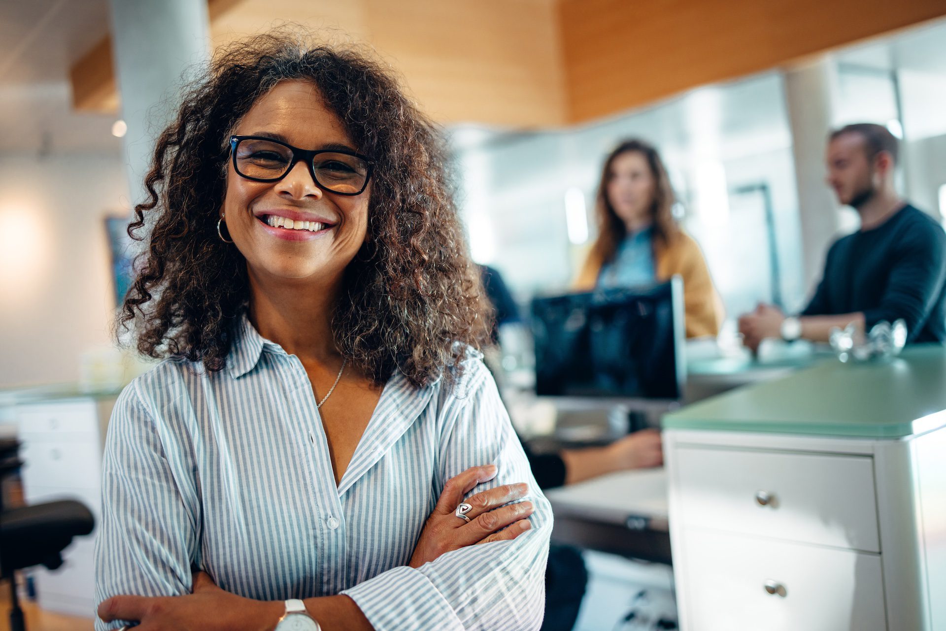 Portrait of a happy woman standing in an office with arms crossed, looking at the camera and smiling, with people at the reception in the background.