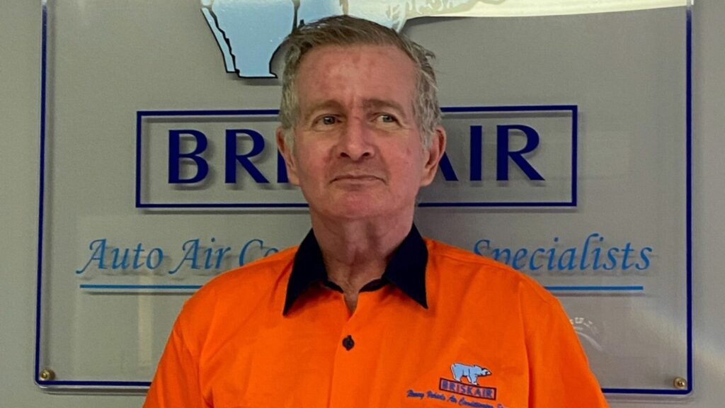 A middle-aged auto air conditioning specialist employee standing in front of the company sign.