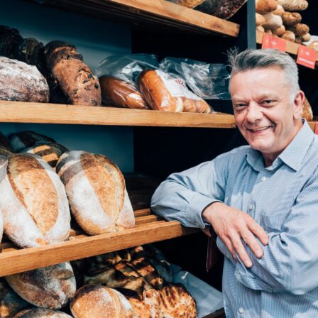 Man smiling, leaning on a shelf of various breads.