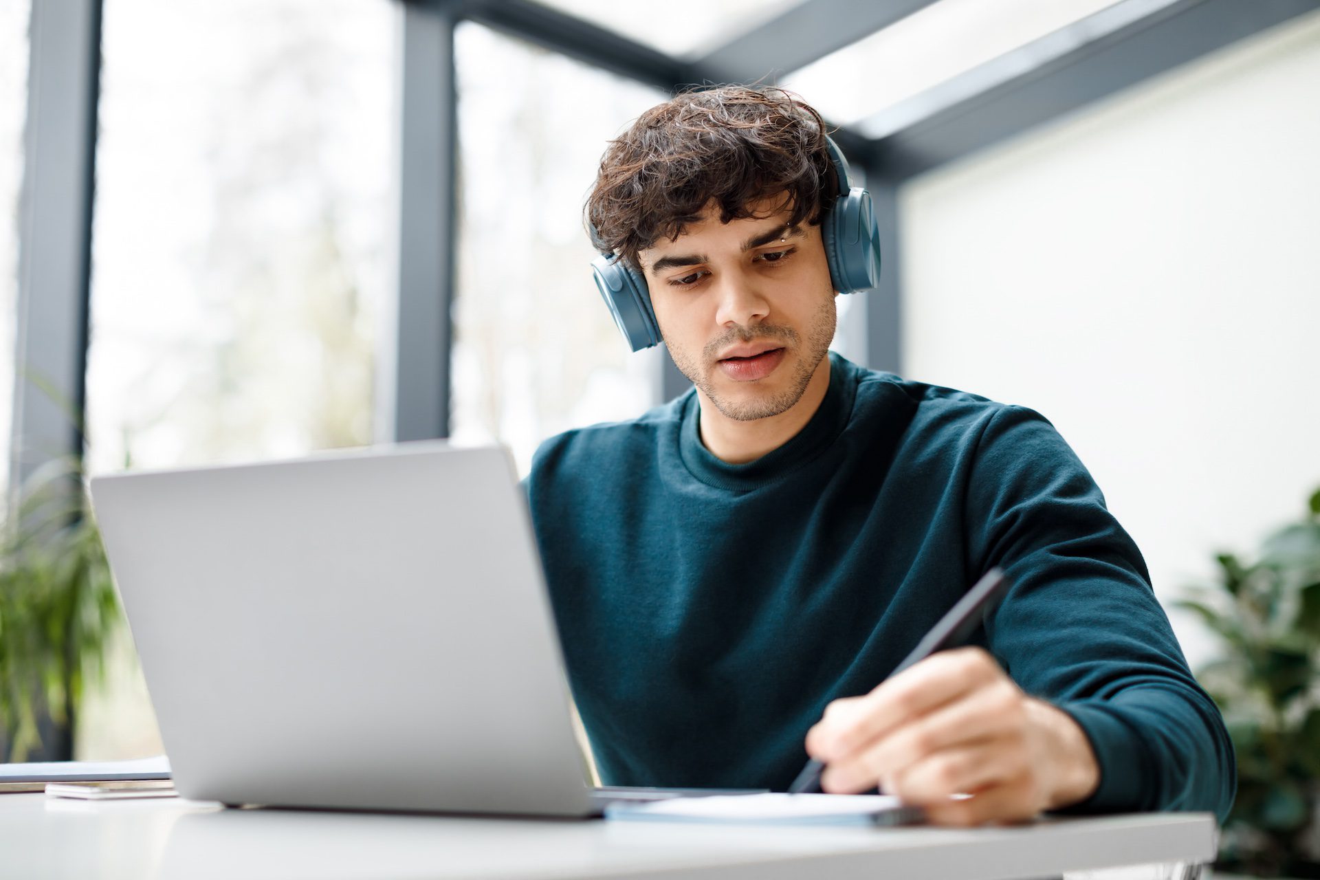 Concentrated European man in headphones studying remotely, watching a video course on a laptop and taking notes.