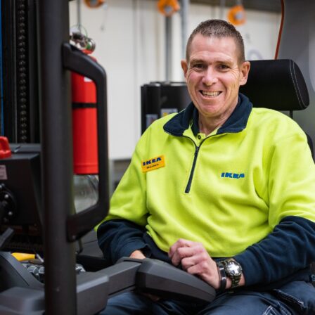 Elder man in an Ikea uniform, smiling while sitting in a forklift.