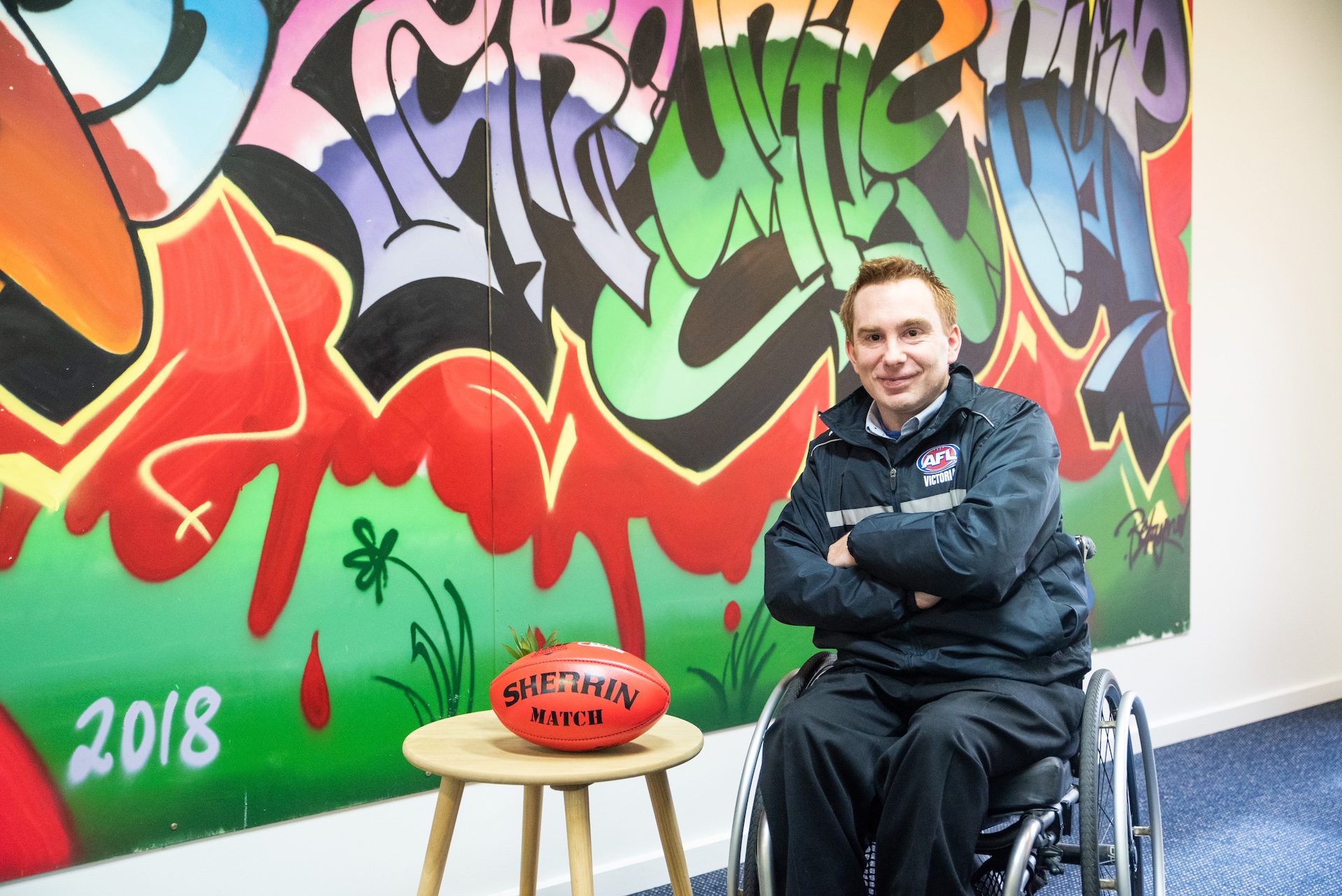 Man in a wheelchair posing by a graffiti wall with a football on a stool.