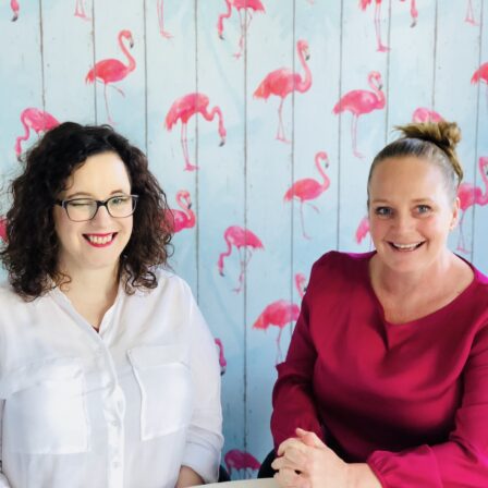 Two women smiling while seated in front of a wall lined with pink flamingo wall paper.