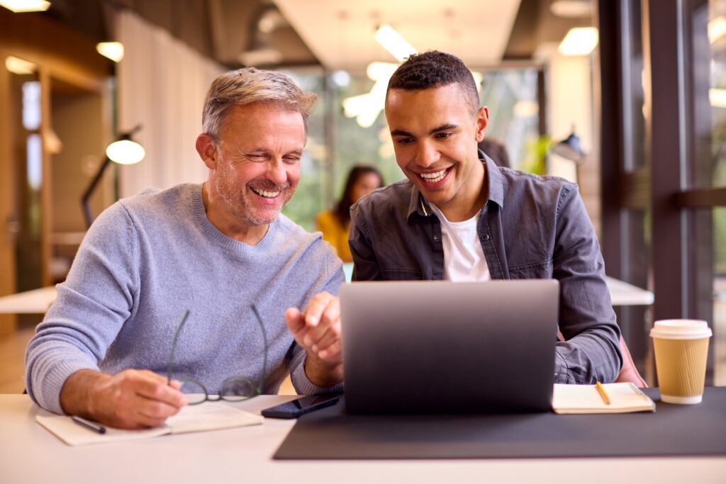 Mature businessman mentoring a younger colleague who is working on a laptop at a desk.