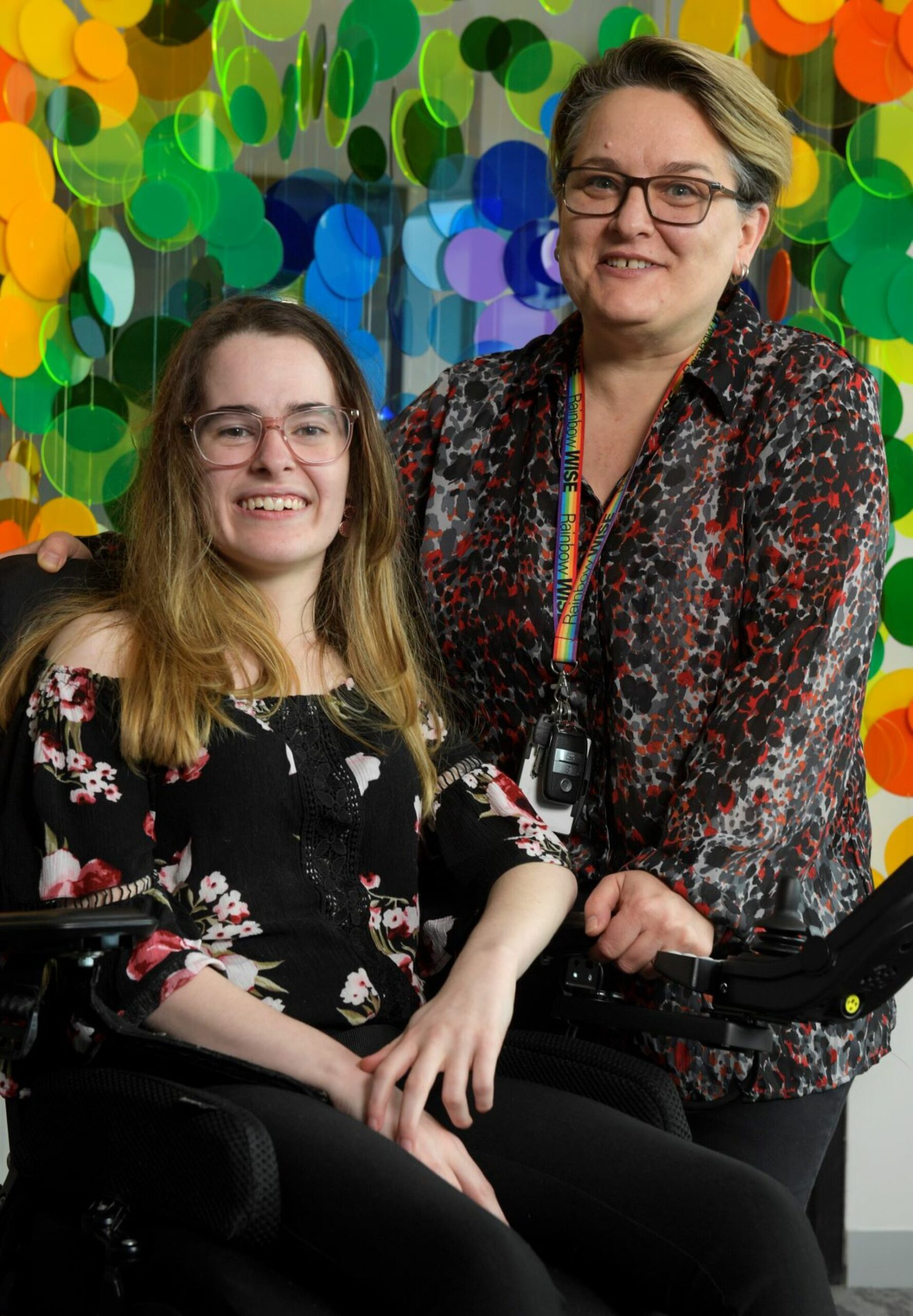 Two women, one in a wheelchair, posing in front of a multicolored background. Ebony