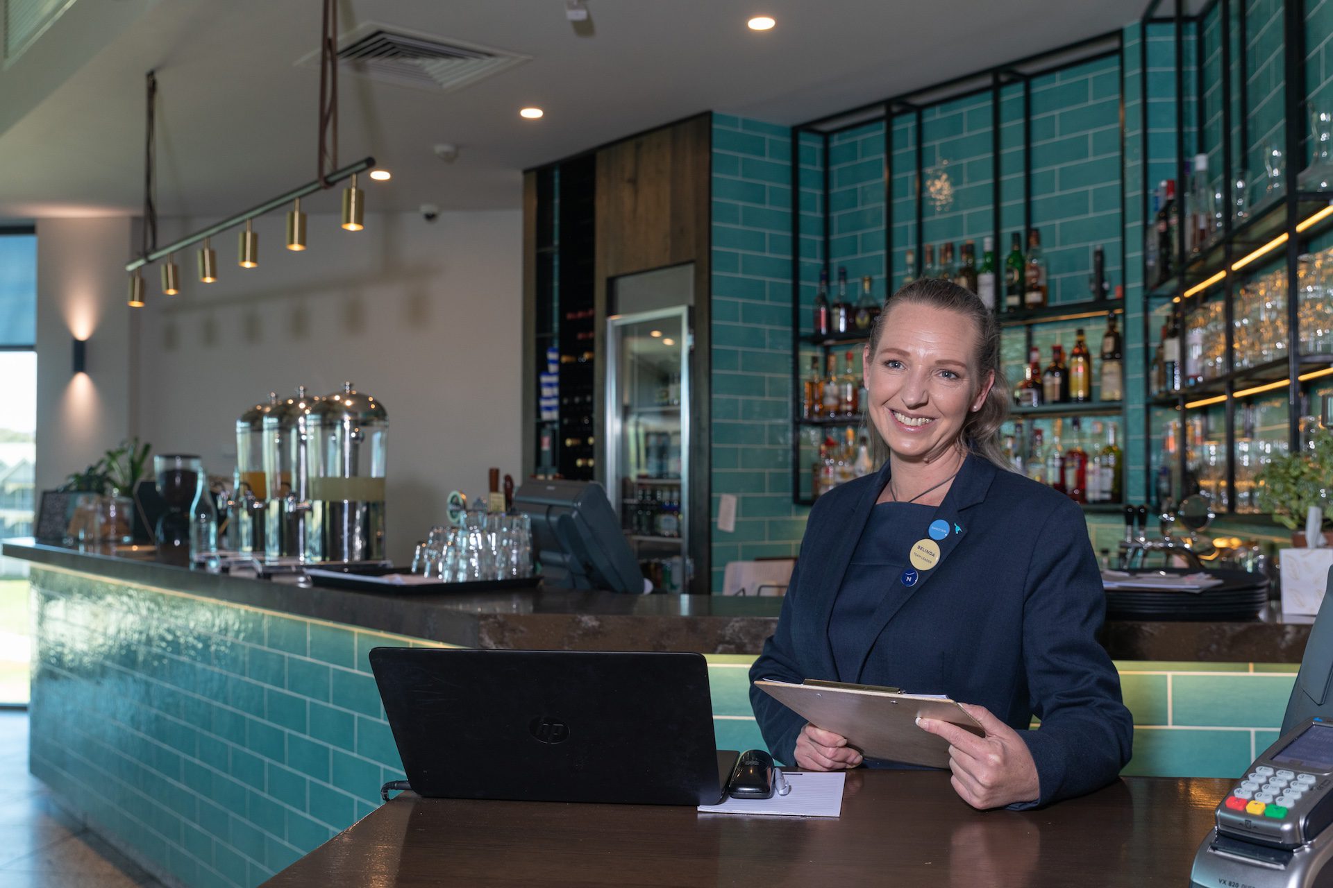 Woman holding a clipboard, smiling while standing in front of a bar counter.