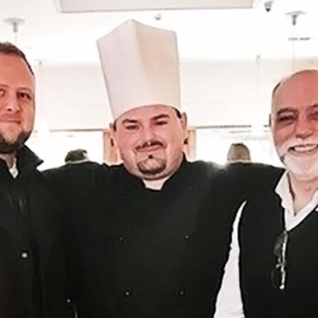 Three smiling men, with the man in the middle wearing a chef's hat.