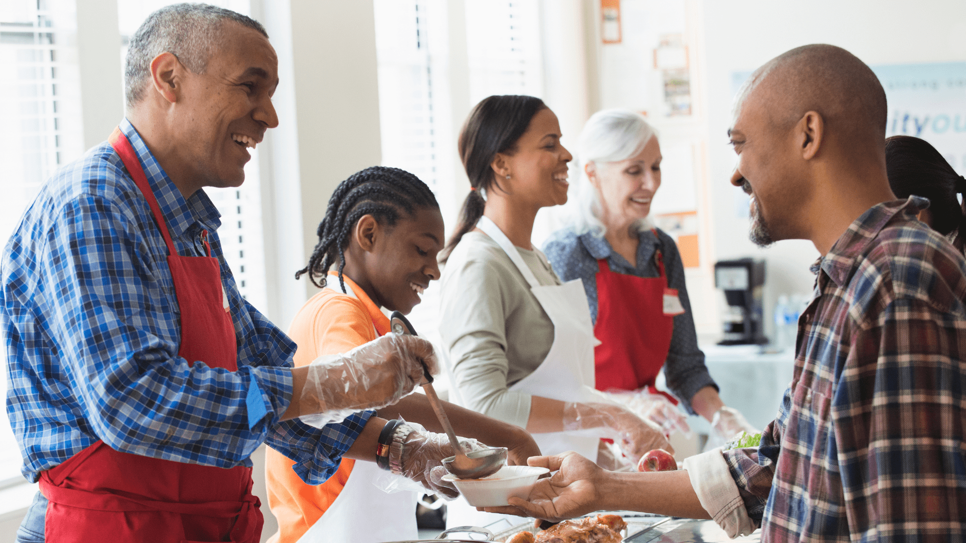 A diverse group of people smiling together, with some serving food from behind a table and another person receiving a meal.