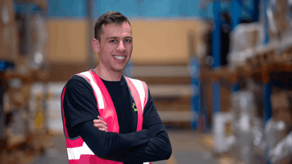 A young man in a warehouse wearing a black shirt and bright pink safety vest smiles with his arms crossed.
