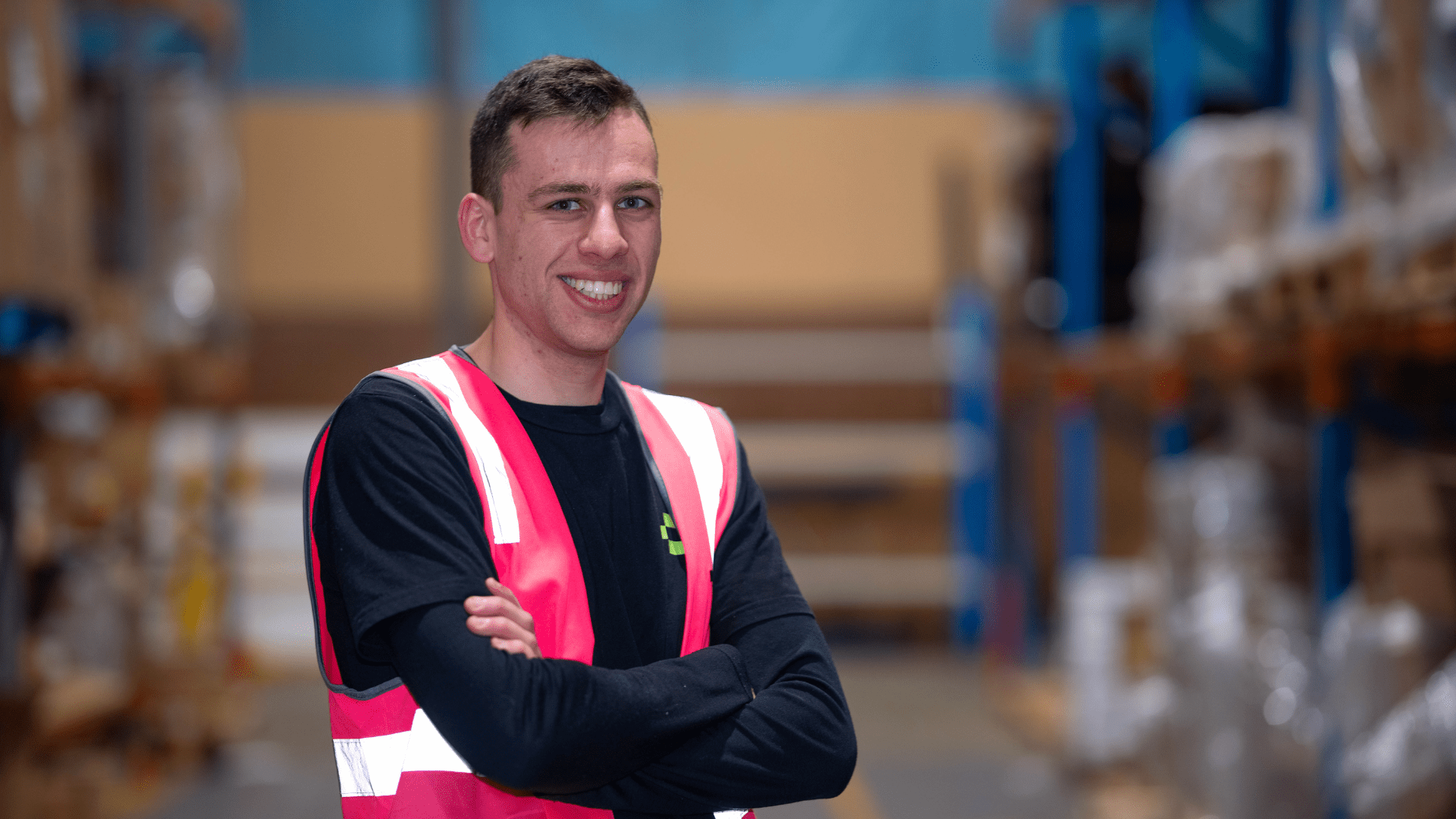 A young man in a warehouse wearing a black shirt and bright pink safety vest smiles with his arms crossed.