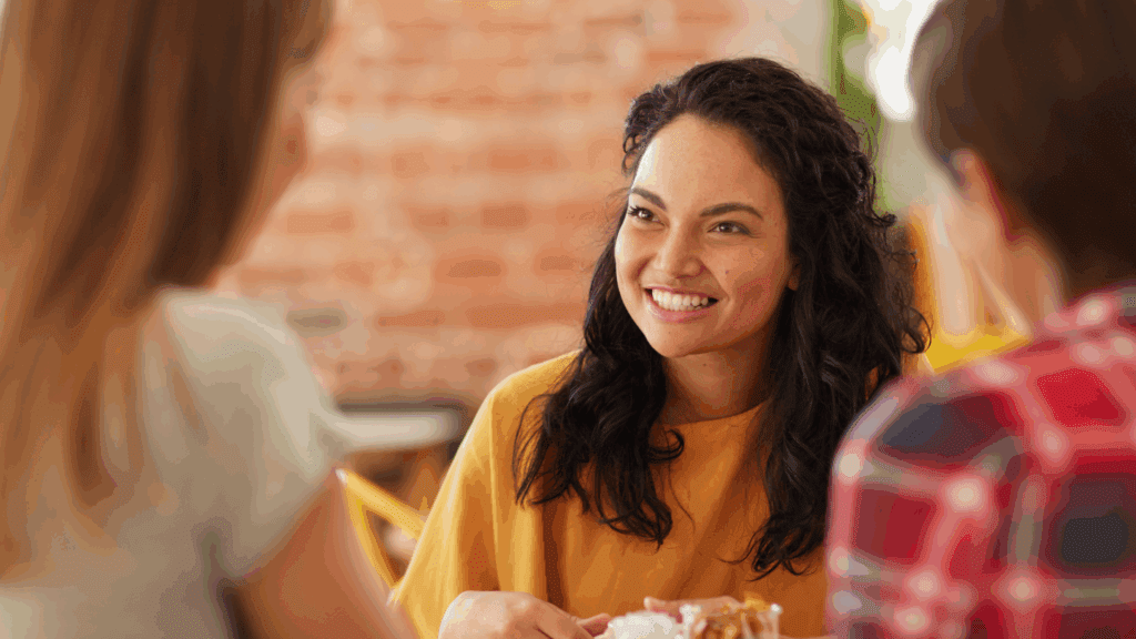 A woman in a mustard-yellow top smiles warmly while talking with two other people in a casual indoor setting