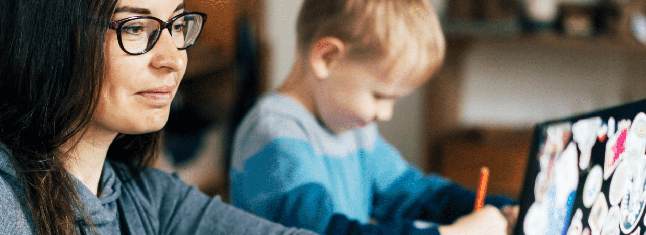 A woman wearing glasses works on a laptop at home while writing notes, with a young child beside her drawing at the table.