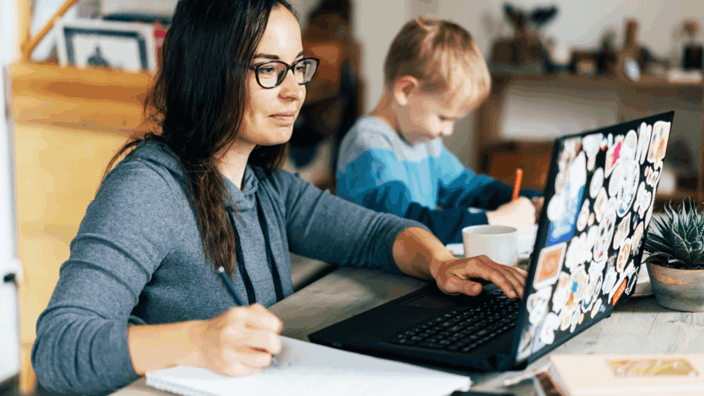 A woman wearing glasses works on a laptop at home while writing notes, with a young child beside her drawing at the table.