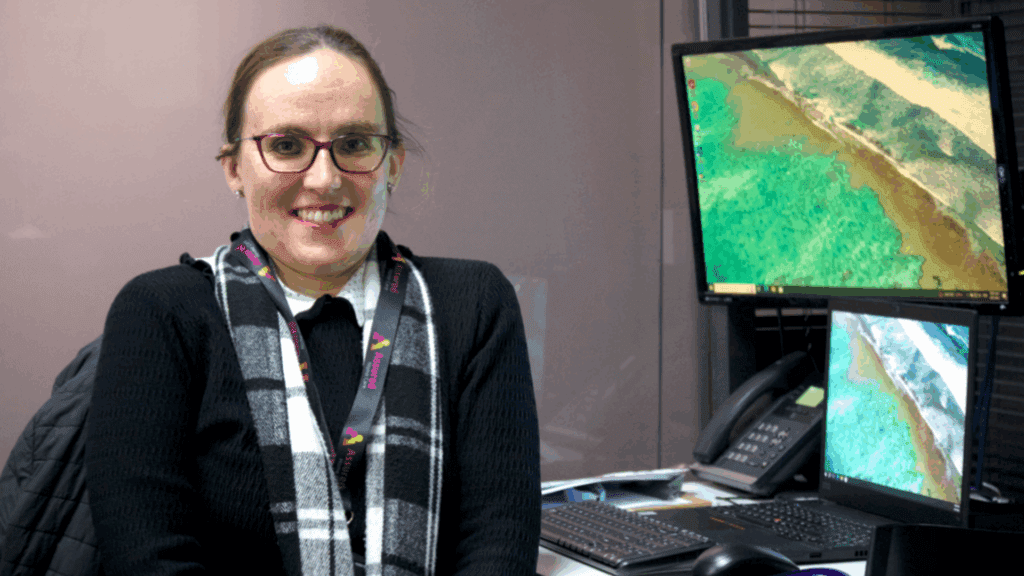 Young woman with glasses and hair tied up, smiling as she sits next to a computer with the screen on