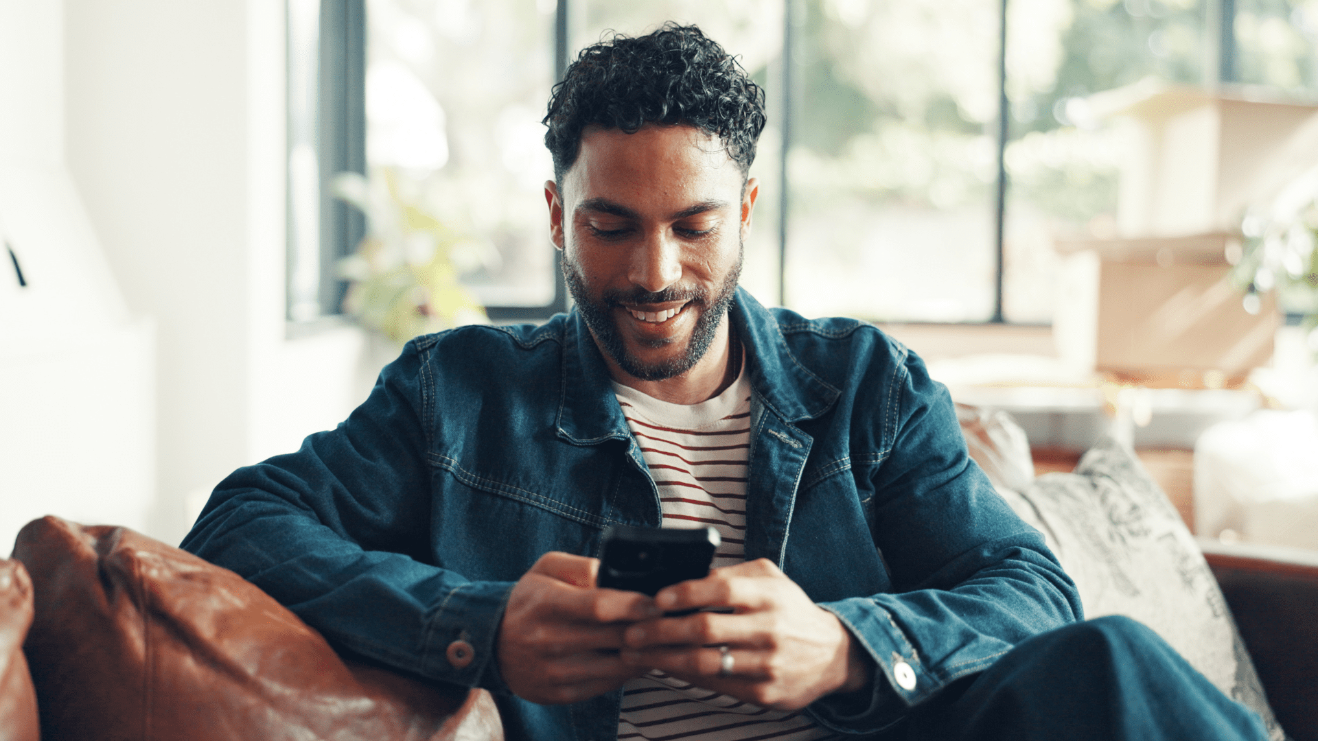 Man sitting on a couch smiling while looking at his phone, wearing a denim jacket and striped shirt in a bright living room