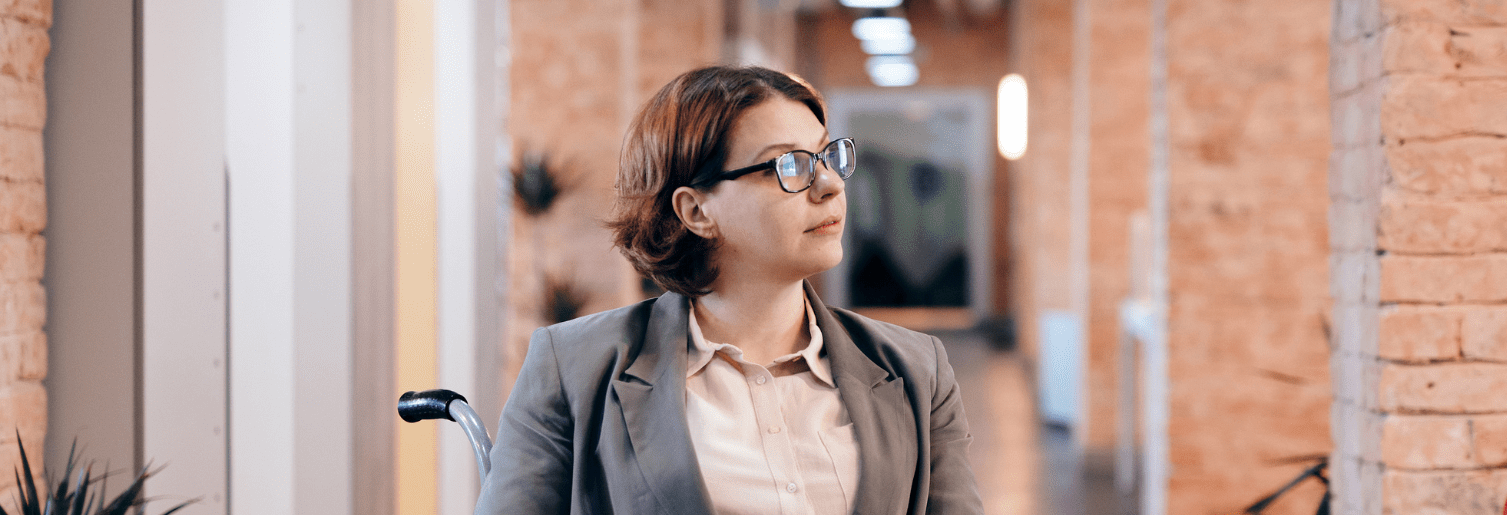 A woman using a wheelchair sits in a bright, modern office hallway, holding a tablet and looking ahead confidently