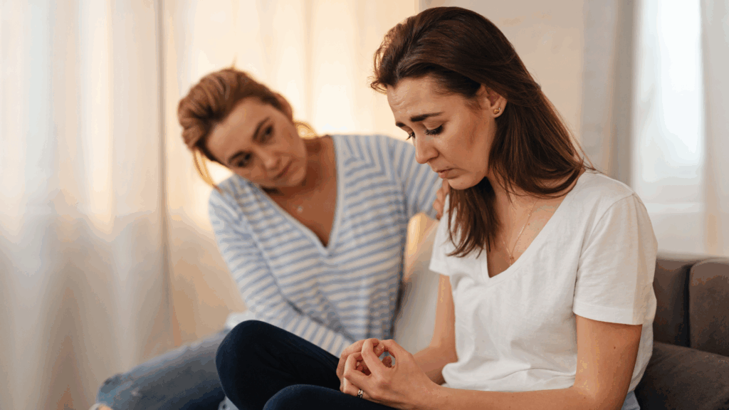 Two women on a couch- one in a white shirt appearing solemn and looking down at her hands, and the other leaning forward to see her reaction with a concerned facial expression.