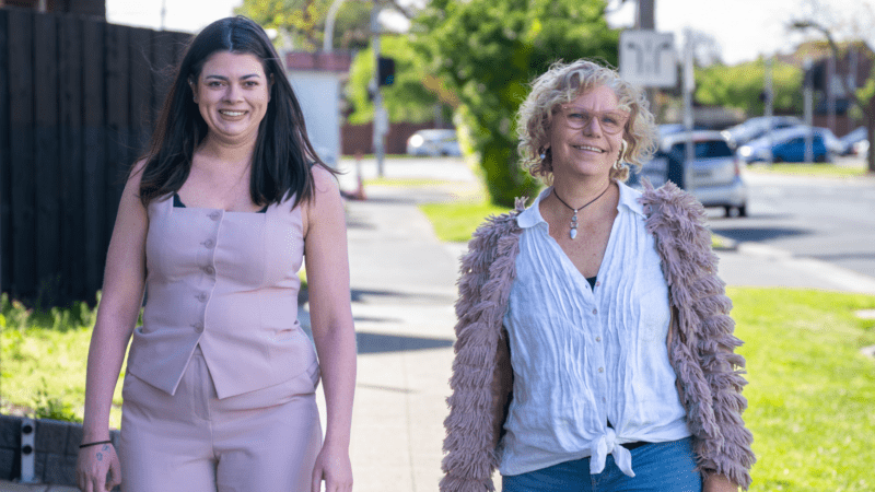 Two smiling women standing on a sunny sidewalk, with trees, grass, and a street in the blurred background