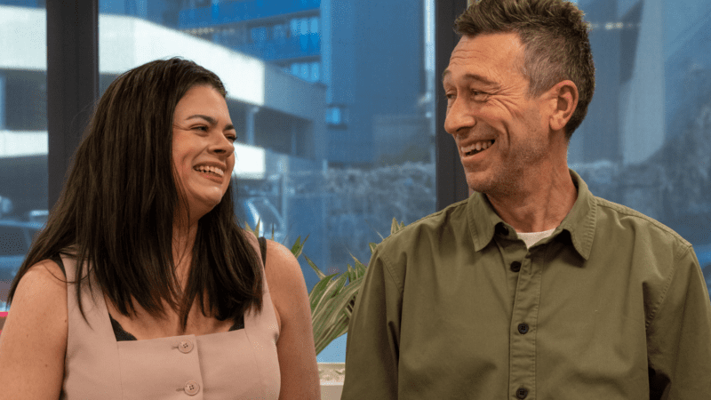 A candid moment of a man and woman laughing together, set against a blurred urban backdrop with a window and greenery