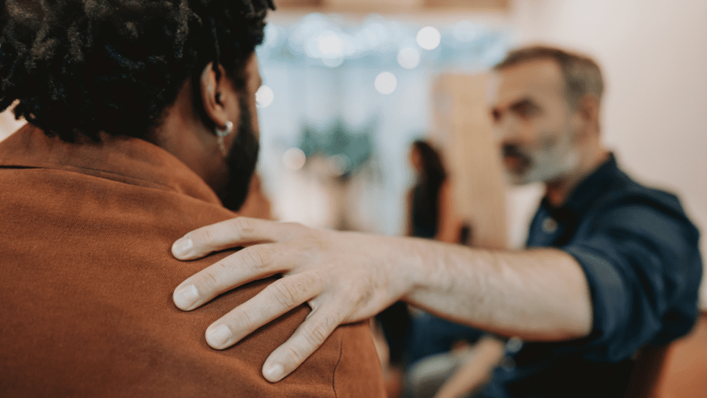 One person places a supportive hand on another person’s shoulder during a workplace conversation.