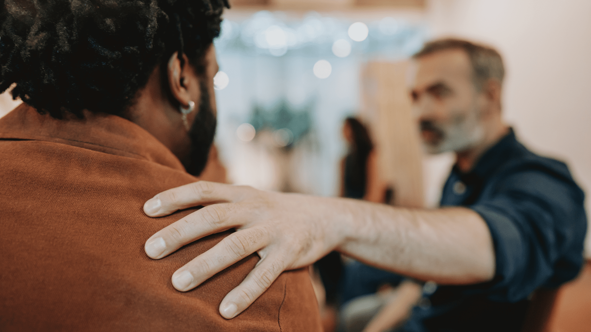 One person places a supportive hand on another person’s shoulder during a workplace conversation.
