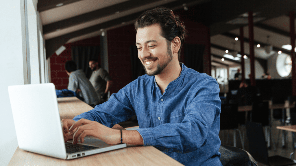 A person using a laptop at a table in a bright, shared workspace.