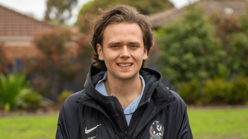 A young man with wavy brown hair and a light stubble, wearing a black jacket with a Nike logo, smiles outdoors with trees and a red-roofed building in the blurred background