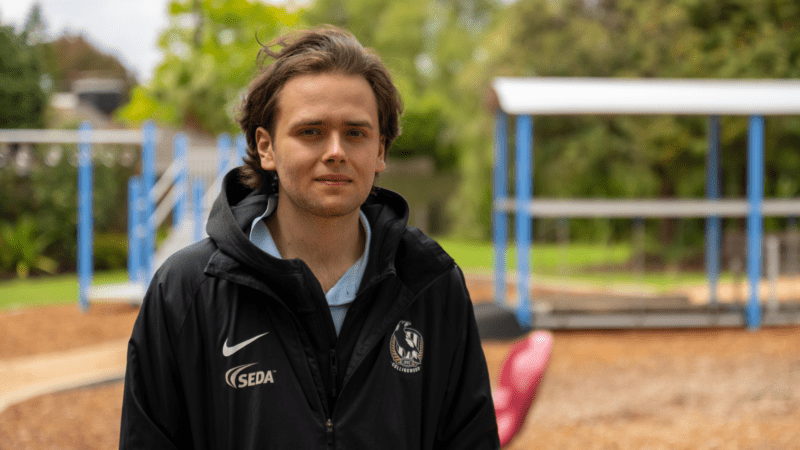 A young man with wavy brown hair and light stubble, wearing a black jacket with "SEDA" and "Collingwood" logos, stands outdoors in a park with playground equipment and trees in the background.