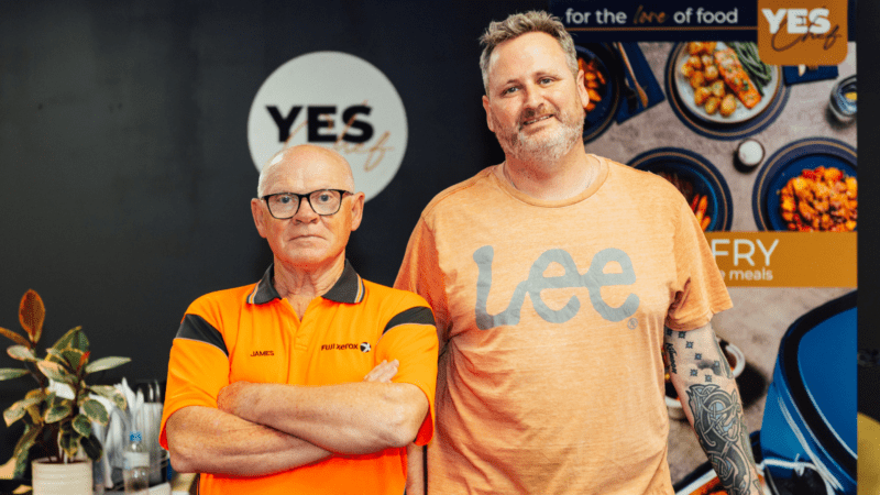 Two men stand in front of promotional materials for 'YES Chef.' The older man on the left, wearing an orange and black polo shirt with 'JAMES' and 'FUJI XEROX,' has his arms crossed. The younger man on the right, in a light orange t-shirt with 'Lee' and a visible tattoo, smiles faintly. The background features a 'YES Chef' logo, food images, and the phrase 'for the love of food.