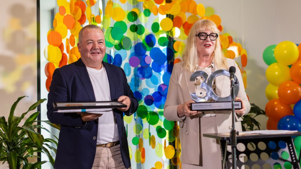 Man wearing white t-shirt under navy blazer holding a framed certificate. He is stood next to a mature blonde woman in a cream coat holding a silver '30' trophy