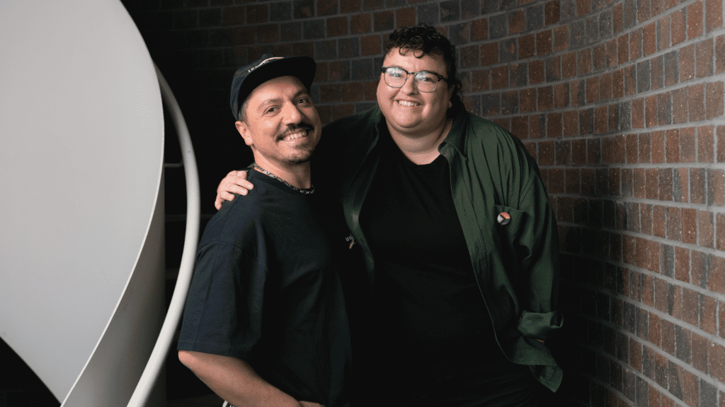 Two people smiling and standing together with their arms around each other beside a brick wall.