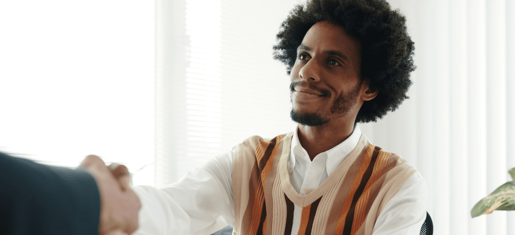 Young man with slight facial hair wearing a knitted vest over a white shirt sitting across the table from someone, shaking their hand