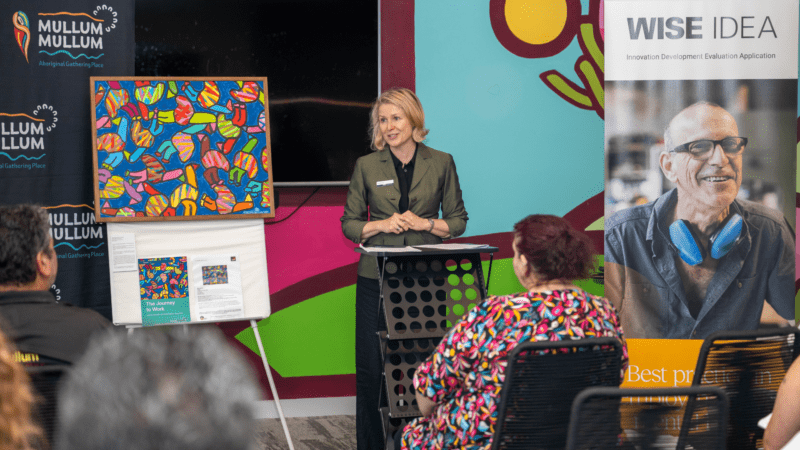 A speaker at a podium during an event, with banners for 'Mullum Mullum Aboriginal Gathering Place' and 'WISE IDEA,' and a colorful abstract painting on display.