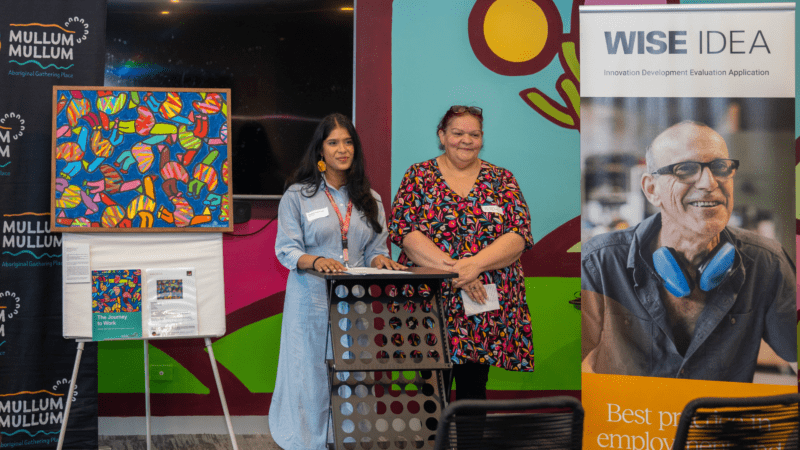 Two women presenting at a podium, flanked by banners for 'Mullum Mullum Aboriginal Gathering Place' and 'WISE IDEA,' with a vibrant abstract painting on an easel