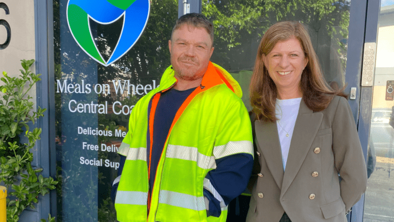 Man wearing a hi-vis jacket over a black t-shirt standing smiling next to a women wearing a grey blazer over a white top and black pants