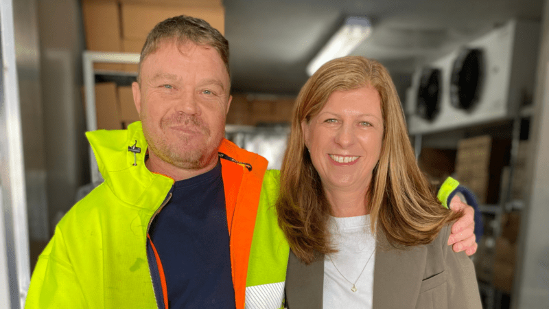Close up photo of man wearing a hi-vis jacket over a black t-shirt standing smiling next to a women wearing a grey blazer over a white top