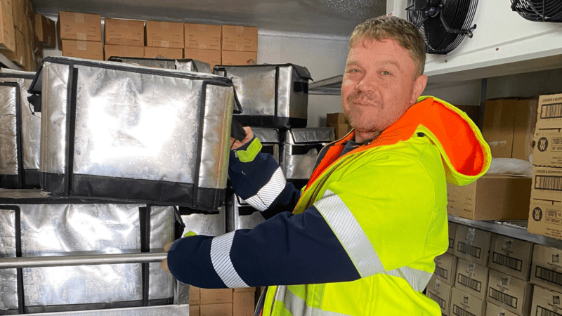 Brendan wearing a hi-vis jacket, standing in a cool room while placing a large silver box on a shelf