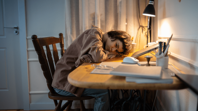 Young person resting their head on a desk beside a laptop, appearing tired or unmotivated while studying or working from home